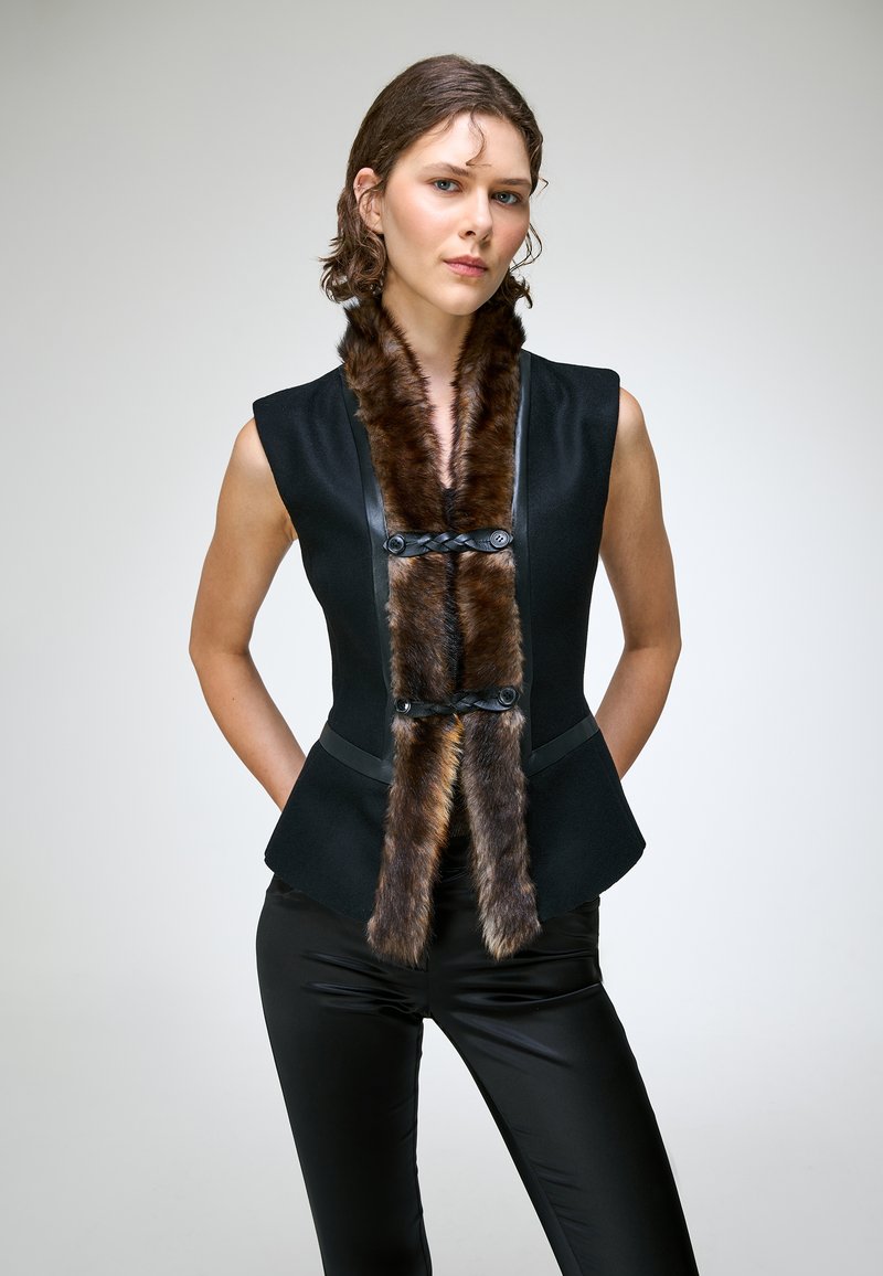 Jeune femme portant un haut noir sans manches avec bordure en fourrure marron et pantalon noir, debout les mains dans le dos sur un fond uni.
