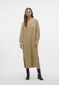 Robe en tricot beige à manches longues, décolleté en V, longueur genoux, avec des fentes sur les côtés et des poignets côtelés, associée à des bottes à talons noires.