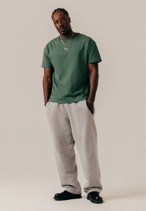 Uomo con i capelli intrecciati, indossa una maglietta verde, pantaloni della tuta grigi, scarpe nere e gioielli d'oro, in piedi con le mani nelle tasche davanti a uno sfondo semplice.