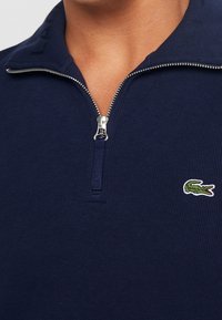 Mörkblå zip-up polo med ribbad textur, featuring en silverdragkedja och en liten grön krokodil-logotyp på bröstet. Klassisk kragsdesign.