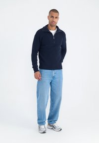 Pulóver de punto azul marino con cuello alto y cremallera en el frente, combinado con vaqueros holgados de color azul claro y zapatillas con detalles en plata.