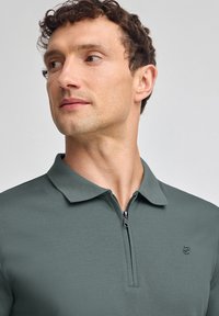 Kortärmad, mörkgrön pikétröja med dragkedja vid kragen och diskret logodetalj på bröstet; slät, lättviktsmaterial.