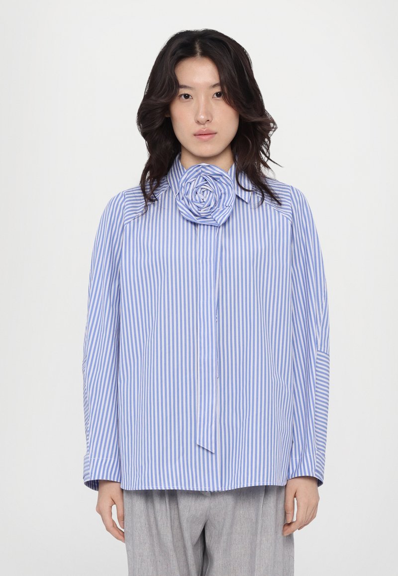Camicia a righe blu e bianca con un accento a forma di rosa sul colletto, con una vestibilità morbida e maniche lunghe. Pantaloni grigi semplici.