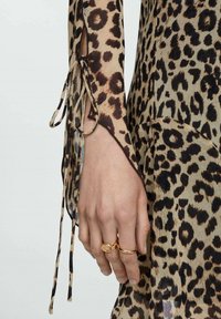Rochie cu imprimeu de leopard, cu mâneci legate la încheietura mâinii, realizată dintr-un material transparent și având un model de culori contrastante, maro și bej. Ineluri aurii pe mână.