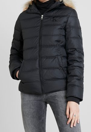 Kvinde iført en sort dunjakke med pelsbelagt hætte, kombineret med grå jeans, hånd i lommen og en synlig lille hjerte-tatovering på fingeren.