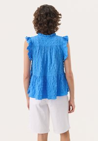 Blaues, strukturiertes Blusenshirt mit Rüschärmeln, gestuftem Design und gerafftem Rücken. Kombiniert mit weißen Shorts, die den lässigen Sommerstil betonen.