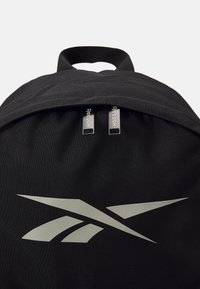 Mochila preta com um tecido liso, alça superior e dois puxadores de zíper prateados com a marca "Reebok", além de um grande gráfico do logotipo.