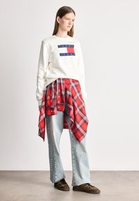 Λευκή μπλούζα με το λογότυπο "TOMMY JEANS", εναλλασσόμενη κόκκινη καρό πουκάμισο δεμένο στη μέση, ανοιχτό γαλάζιο παντελόνι σε φαρδιά γραμμή, και μαύρα sneakers με λεοπάρ μοτίβο.