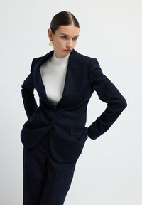 Námořnický blazer s pruhy a klopami, se dvěma předními kapsami, nošený přes bílý žebrovaný rolák, doplněný o odpovídající kalhoty.