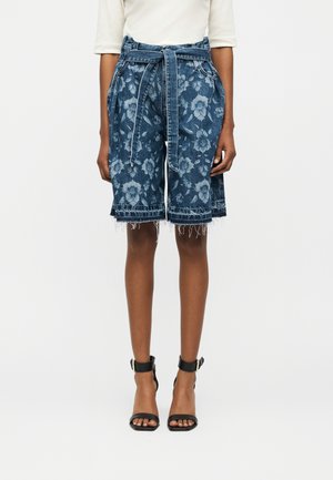 Naine, kes kannab kõrge vöökohaga teksapükse lukuäärte ja lillemustriga ning roheliste sangadega mustade pahkluusussidega sandaale.