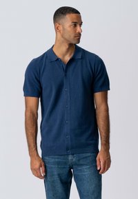 Marineblå kortærmet polo lavet af tekstureret strikket stof, med knaplukning foran, krave og en behagelig pasform. Kombineret med blå jeans.