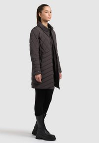 Woman wearing a mid-length dark quilted coat, black leggings, and black lace-up chunky boots standing against a plain light background.