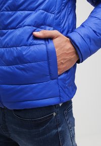 Blue quilted puffer jacket with a zippered front, side pockets, and a smooth texture. Fits closely around the body with a slightly shiny finish.