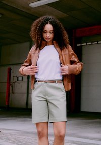 Kvinne med krøllete hår som justerer brun skinnjakke, iført hvit t-skjorte, beige shorts, og står i et urbant miljø.