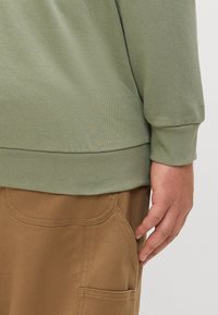 Olijfgroene blouse met lange mouwen en geribbelde manchetten, gedragen over beige stofbroek met een zak en gedetailleerde stiksels. Gladde textuur zichtbaar.