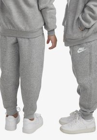 Tricou și pantaloni de trening gri din material moale, cu manșete elastice. Logo-ul Nike alb pe pantaloni. Adidași albi cu șireturi.