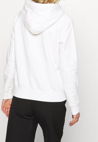 Sudadera blanca con capucha y corte relajado. Presenta puños y dobladillo de canalé, textura suave, y una costura visible en la parte trasera.