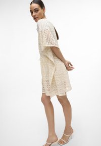 Abito in pizzo color crema con maniche corte, caratterizzato da un motivo a zigzag, vestibilità ampia e vita cinchata. Abbinato a sandali leggeri con cinturini.