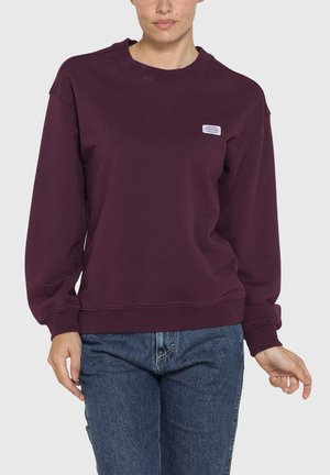 Persona che indossa una felpa girocollo viola scuro con un piccolo logo bianco sul petto e jeans blu, in piedi su uno sfondo semplice.