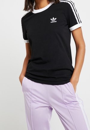 Person trägt ein schwarzes Adidas-T-Shirt mit weißen Streifen an den Ärmeln und hellviolette Hose, Hände in den Taschen, vor einem einfarbigen Hintergrund.