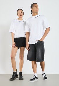 Hvite kortermede t-skjorter med svart piggtrådsbroderi, kombinert med et svart plisserte skjørt og denimshorts. Sorte støvler og sneakers brukes.