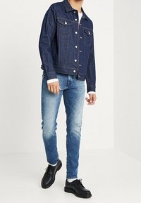 Denim jacket in dark blue, featuring two chest pockets and silver buttons. Paired with light blue distressed skinny jeans and black shoes.