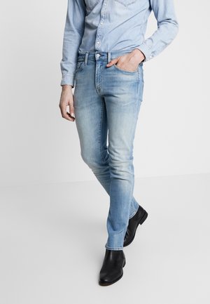 Personne portant un jean en denim bleu clair, une chemise en denim clair et des bottines noires, debout avec une main dans la poche et une jambe croisée vers l'avant.
