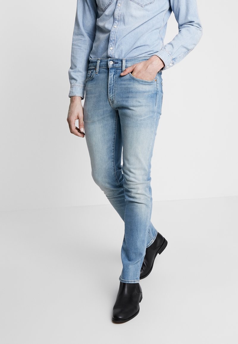 Personne portant un jean en denim bleu clair, une chemise en denim clair et des bottines noires, debout avec une main dans la poche et une jambe croisée vers l'avant.