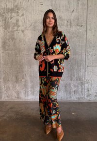 Γυναίκα στέκεται μπροστά σε σκυρόδετο τοίχο φορώντας μαύρη ζακέτα με λουλουδάτο μοτίβο και αντίστοιχο παντελόνι, με καφέ μυτερά παπούτσια.