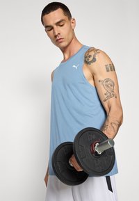 Sports tanktop i lyseblå stof med et hvidt logo. Personen holder en sort håndvægt med metalliske accenter. Strukturerede tatoveringer på armen.