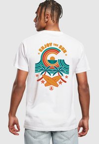 Hvit bomulls t-skjorte med en fargerik grafikk av bølger, sol, palmer og surfebrett. Tekst "NYT SOLEN" i fet oransje og teal.