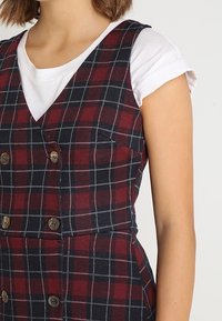 Karo-Kleid mit burgunderfarbenem und marineblauem Muster, das über einem weißen Hemd getragen wird. Es hat einen V-Ausschnitt, Knopfdetails und besteht aus Baumwollstoff.
