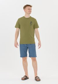 Olivia vihreä lyhythihainen t-paita, jossa on palmuprintti, yhdistettynä sinisiin käärittyihin shortseihin ja ruskeisiin sandaaleihin. Malli seisoo kädet rentoina.