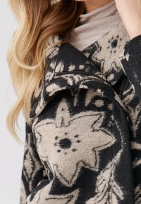 Abrigo de mezcla de lana con un patrón floral en negro y beige, cuello asimétrico y detalle de bolsillo con cremallera en el costado.