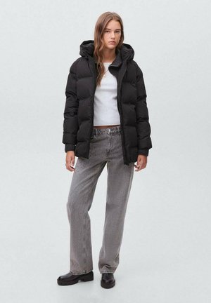 Svart pufferjacka med huva, kviltat mönster och ribbade ärmslut, bärs över en vit skjorta, kombinerad med grå löst sittande jeans och svarta stövlar.