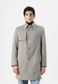 Grau Wollmantel mit hohem Kragen, vorderen Knöpfen und seitlichen Taschen. Besitzt eine Klappe über einer Schulter und eine gerade Silhouette.