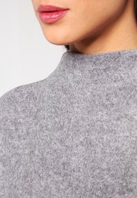 Maglione grigio a collo alto realizzato in un morbido tessuto testurizzato. La foto in primo piano mostra la superficie liscia e le sottili variazioni di colore. Nessun hardware o motivo visibile.