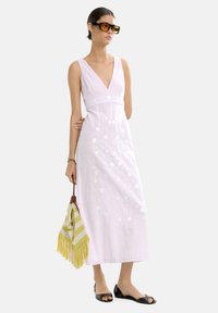 Długa różowa sukienka z dekoltem w serek, haftem kwiatowym i dopasowaną talią. Czarne płaskie sandały i torba w paski w kolorze żółtym i białym z frędzlami.