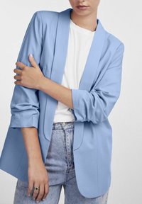 Blazer azzurro chiaro con maniche lunghe, rever a taglio e tessuto morbido. Indossato sopra una t-shirt bianca e abbinato a jeans in denim a vita alta.
