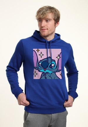 Joven con sudadera azul con capucha con un personaje de alienígena de dibujos animados y estrellas en el frente, de pie con las manos cerca de los bolsillos, mirando hacia la derecha.