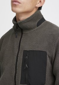 Fleecejack in olijfgroen met een hoge kraag, zwarte stofaccenten, een ritszak op de borst en een gestructureerd oppervlak.