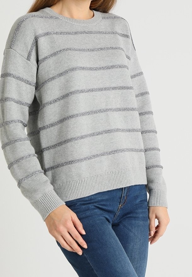 Femme portant un pull gris clair avec des rayures horizontales gris foncé et un jean bleu, debout devant un fond clair uni.