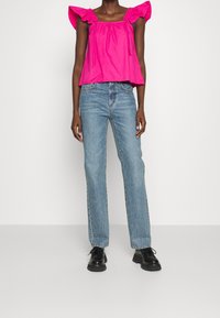 Persona vestida con una blusa de mangas rizadas de color rosa brillante, jeans rectos de mezclilla azul claro y zapatos negros de cordones gruesos sobre un fondo blanco.