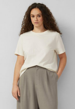 Kobieta o kręconych włosach, ubrana w luźną, białą koszulę z krótkim rękawem, wpuszczoną do wysokowyższych szarych spodni, stojąca na jednolitym tle.