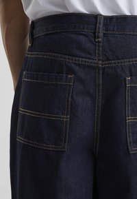 Tummat denimhousut, joissa on korkea vyötärö, suuret takataskut ja kontrastiväriset keltaiset tikkaukset. Sileä pinta ja jämäkkä istuvuus.