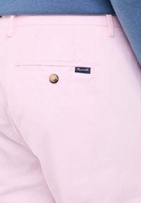 Lichtroze katoenen chinos hebben een achterzak met een marineblauw etiket, messing knopen en een gladde textuur. Toegankelijke snit zichtbaar.