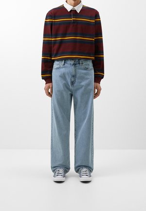 Person trägt ein bordeauxrot gestreiftes Rugbyshirt mit langen Ärmeln, hellblaue locker sitzende Jeans und graue Sneakers und steht vor einem einfarbigen Hintergrund.