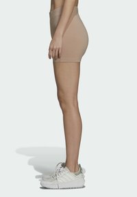 Shorts stretch beiges avec une taille haute, présentant une texture lisse. Portés avec des baskets blanches et vert clair. Profil latéral visible.