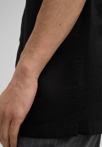 Schwarzes T-Shirt mit einer glatten Textur, seitlichen Nahtdetails und einem lockeren Schnitt, kombiniert mit grauen Nadelstreifenhosen. Keine sichtbaren Logos.