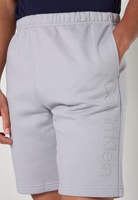 Pantaloncini di cotone grigio chiaro con vita elasticizzata, caratterizzati da tasche laterali e dal logo "Calvin Klein" a rilievo sulla gamba sinistra.